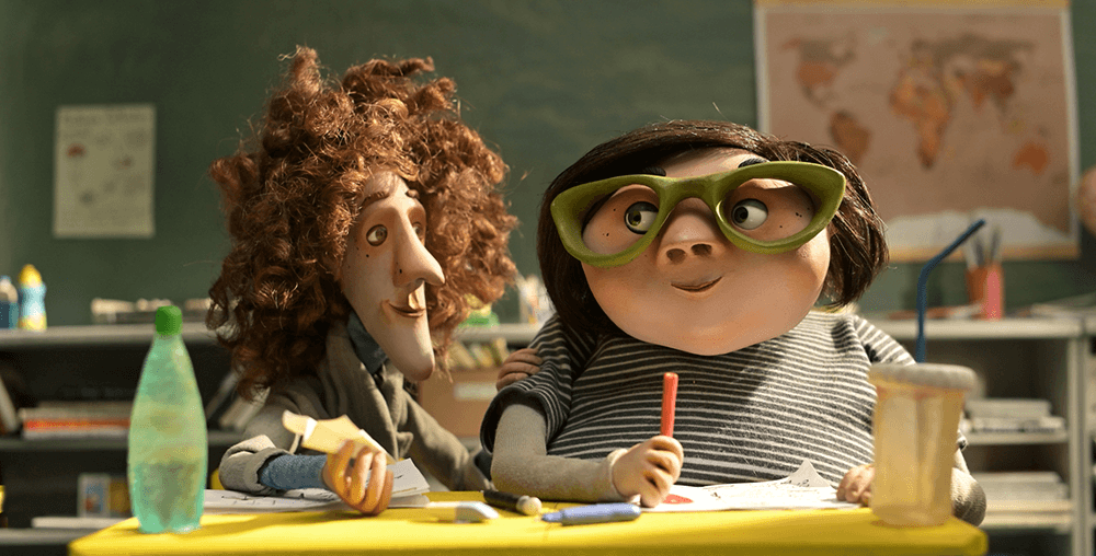 Two animated puppets from a stop-motion film sit at a school desk smiling slightly