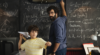 a young boy aims a paper airplane while a man writing on a large chalkboard behind him